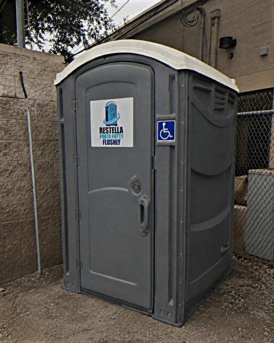 Grey accessible portable restroom unit standing against a block wall on a work site