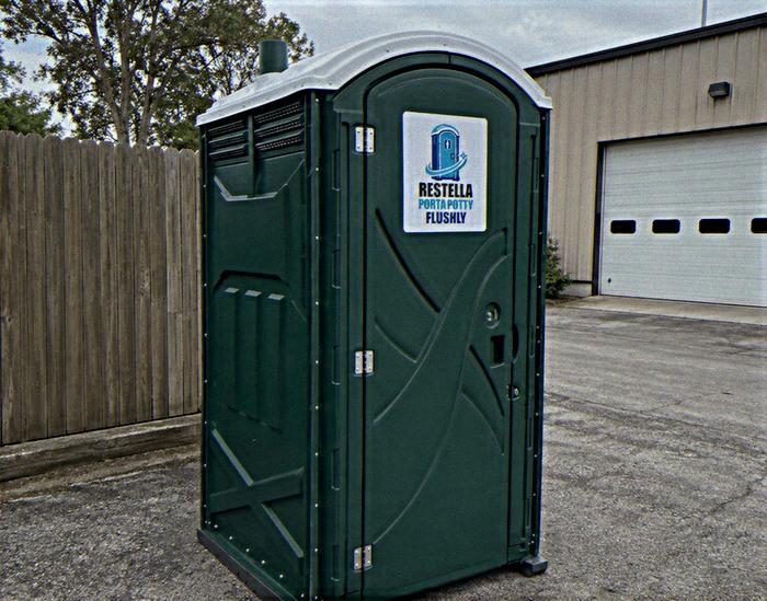 A single green standard portable toilet unit parked on a paved lot beside a work building